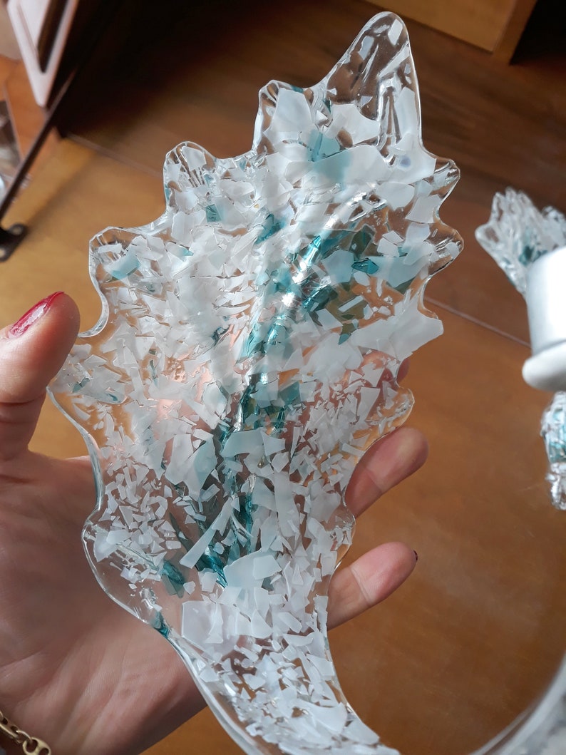 K&ouml;nnte beinhalten: Ein dekoratives St&uuml;ck aus klarem Glas in Blattform, gef&uuml;llt mit wei&szlig;en und t&uuml;rkisblauen Scherben. Das Glas wird in einer Hand gehalten, wodurch sein kompliziertes Design und seine Textur hervorgehoben werden.