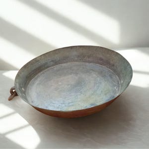 May include: A shallow, round metal basin with a weathered, tarnished interior surface showing shades of grey, blue, and green. The exterior of the basin features a warm copper tone, and a small, curved metal handle is attached to the side rim.