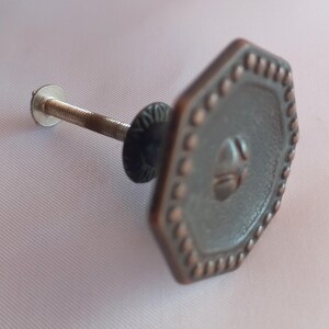 May include: An octagonal cabinet knob with a dark metallic finish and a copper-toned beaded border. The center features a raised geometric design. A threaded metal bolt extends from the back, accompanied by a small circular washer and nut.