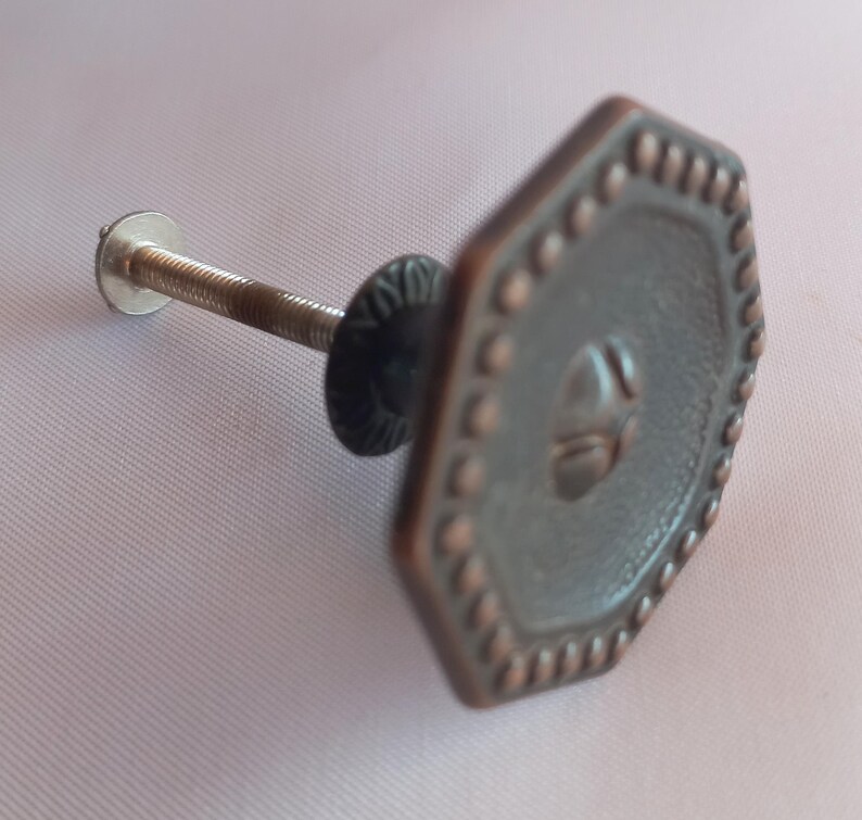 May include: An octagonal cabinet knob with a dark metallic finish and a copper-toned beaded border. The center features a raised geometric design. A threaded metal bolt extends from the back, accompanied by a small circular washer and nut.