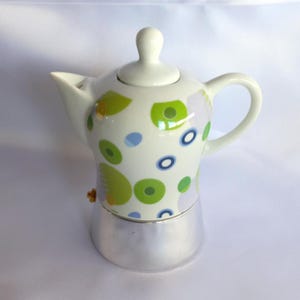 Puede incluir: Una cafetera italiana de cerámica blanca con un diseño de círculos y óvalos verdes y azules. La mitad inferior es de metal plateado, con un asa y tapa blancos.