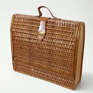 May include: A brown wicker handbag with a leather handle and clasp. The bag has a woven pattern and a curved top. The leather clasp is secured with a gold-coloured metal button.