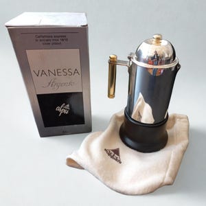 Könnte beinhalten: Eine silberbeschichtete Espressomaschine mit schwarzem Sockel und goldenem Griff, neben der Verpackung. Auf der Schachtel steht "VANESSA Argento" und "Caffettiera express in acciaio inox 18/10 silver plated."