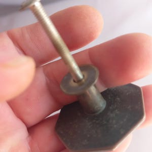 May include: A metallic cabinet knob with an octagonal face and a dark, weathered finish. The knob features a long, threaded metal bolt extending from the center of the back, designed for attachment to furniture or cabinetry doors and drawers.