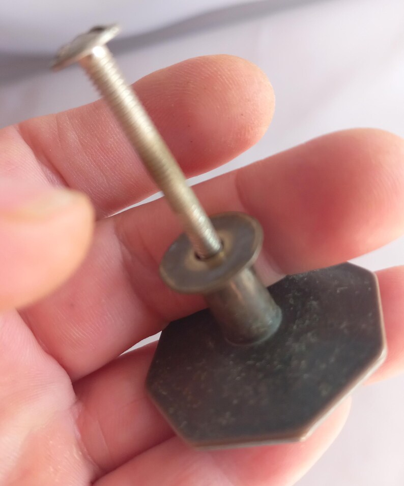 May include: A metallic cabinet knob with an octagonal face and a dark, weathered finish. The knob features a long, threaded metal bolt extending from the center of the back, designed for attachment to furniture or cabinetry doors and drawers.