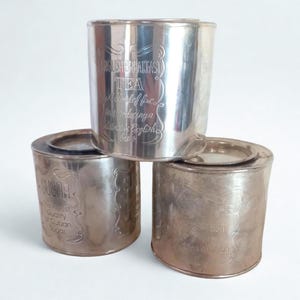Op de afbeelding: Drie zilverkleurige blikken op elkaar gestapeld. Op het bovenste blik staat in een elegant lettertype "English Breakfast Tea". Op het middelste blik staat "Sugar" en op het onderste blik "Tea". De blikken hebben een vintage esthetiek.