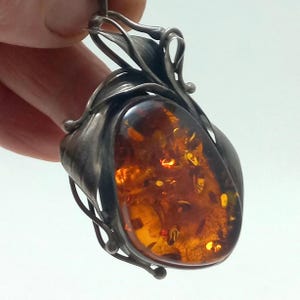 Può includere: Ciondolo in argento con una grande pietra d'ambra ovale. L'ambra è di un colore arancione caldo e traslucido con inclusioni. Il ciondolo presenta un design argentato dettagliato a forma di foglia attorno all'ambra, con un anello per una catena.