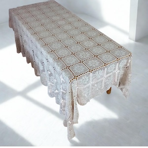 May include: A cream-coloured crochet tablecloth, rectangular in shape. It covers a wooden table, displaying a detailed square pattern. The edges have a scalloped design. The tablecloth's neutral colour makes it suitable for various decor styles. The table is approximately 180cm long.