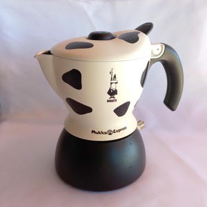 Pode incluir: Uma cafeteira Bialetti Mukka Express com a parte superior creme com manchas pretas de vaca, uma base preta e uma pega preta. O logótipo Bialetti e o texto Mukka Express estão impressos na parte frontal da unidade.