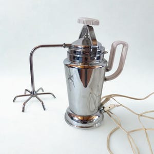 Può includere: Frullatore elettrico vintage cromato con manico e parte superiore grigio chiaro. Il frullatore ha un braccio lungo e curvo con un accessorio di miscelazione a forma di stella. Un cavo di alimentazione con spina bianca esce dalla base.