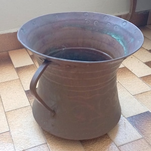 May include: A copper-colored metal pitcher with a handle. The pitcher has a wide, flared rim and a bulbous body. The handle is attached to the side of the pitcher. The metal has a slightly aged appearance.