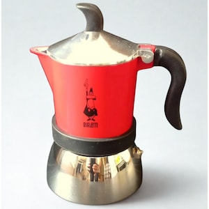 Op de afbeelding: Een rode en zilveren espressomaker voor op het fornuis met een zwarte handgreep en deksel. De rode behuizing heeft een zwarte afbeelding en het woord "BIALETTI". De espressomaker is ontworpen voor gebruik op het fornuis en is gemaakt van metaal.