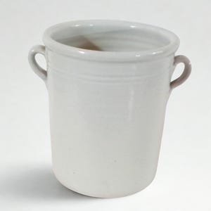 Op de afbeelding: Een cilindervormige keramische pot met een glanzende gebroken witte afwerking, voorzien van twee kleine, ronde handvatten aan weerszijden bij de rand. De houder heeft een glad, uniform oppervlak en een licht uitlopende bovenrand voor gebruik.