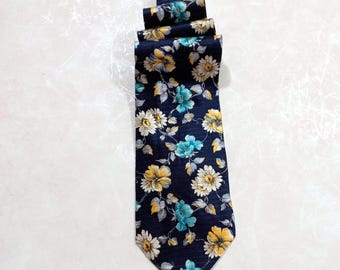 May include: A navy blue necktie with a floral pattern. The tie features a repeating design of yellow, white, and turquoise flowers with green leaves. The tie is made of a smooth fabric and is suitable for formal or semi-formal occasions.