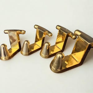 May include: Four gold-colored metal hooks arranged in a row. Each hook has a J-shaped design with a rounded top and a conical base. The hooks are made of a shiny, reflective material.