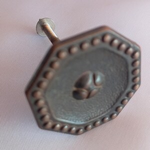 May include: An octagonal cabinet knob with a dark bronze finish, featuring a beaded border and a central raised circular motif. The knob includes a threaded metal bolt extending from the back for attachment to furniture or cabinetry surfaces.