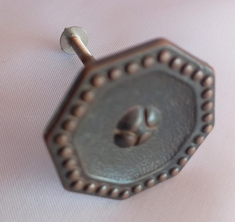 May include: An octagonal cabinet knob with a dark bronze finish, featuring a beaded border and a central raised circular motif. The knob includes a threaded metal bolt extending from the back for attachment to furniture or cabinetry surfaces.