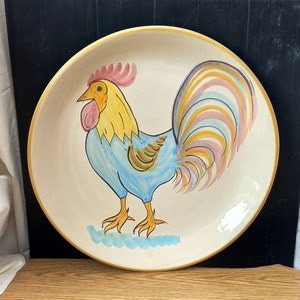 May include: A large, round ceramic plate with a hand-painted rooster design. The rooster features a yellow head, blue body, and a colorful tail with pink, yellow, and blue accents. The plate has a cream-colored background with a yellow rim.