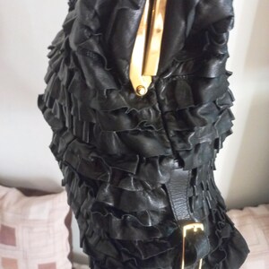 May include: Black leather handbag with a ruffled design. The bag has a gold-coloured clasp and buckle. The texture is soft and the design is eye-catching. The bag is a stylish accessory.