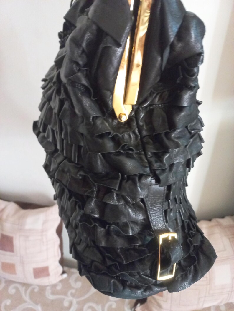 May include: Black leather handbag with a ruffled design. The bag has a gold-coloured clasp and buckle. The texture is soft and the design is eye-catching. The bag is a stylish accessory.