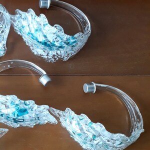 K&ouml;nnte beinhalten: Klare Glasservierschalen mit wellenf&ouml;rmigem Design. Jede Schale hat einen gebogenen Griff mit einem silberfarbenen Metallende. Das Innere der Schalen ist mit kleinen, blau gef&auml;rbten Glasst&uuml;cken gef&uuml;llt.