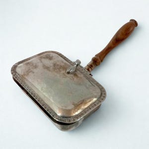 Puede incluir: Un recogedor de migas vintage plateado con mango de madera. El recogedor rectangular tiene un borde decorativo y una tapa con bisagras. El mango es de color marrón oscuro. Está diseñado para recoger migas de la mesa.