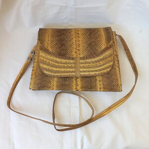 May include: A square, brown and yellow snakeskin crossbody bag with a flap closure. The bag has a long, thin strap and a striped design on the flap. The bag is made of a snakeskin material.