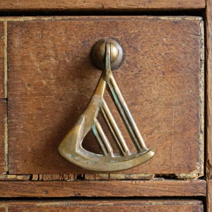May include: A brass drawer pull with a triangular, fan-like shape featuring three vertical slats. The metal has a weathered, aged patina with visible green oxidation in the crevices, mounted onto a flat, dark brown wooden drawer front.
