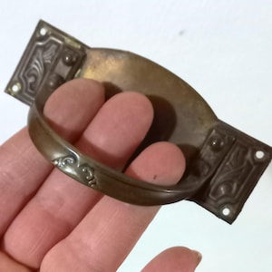 Può includere: Maniglia per cassetto antica color bronzo con manico curvo e piastre terminali rettangolari decorative. Le piastre terminali presentano disegni floreali in rilievo e fori di montaggio. Un pezzo di ferramenta vintage.