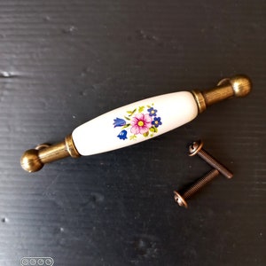 Peut inclure: Une poignée de meuble en céramique blanche ornée d'un motif floral rose, bleu et jaune. Elle est dotée d'extrémités en métal couleur laiton et est fournie avec deux vis. Cet accessoire est décoratif pour les meubles.