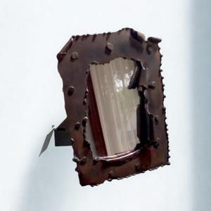 May include: A copper-coloured metal mirror with a rectangular shape and a textured, irregular frame. The mirror is mounted on a small, angled metal stand. The mirror is designed to be wall-mounted.