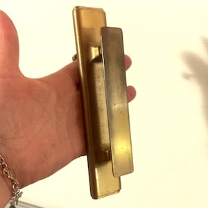 Op de afbeelding: Een gouden deurknop met een rechthoekige achterplaat en een lange, smalle handgreep.