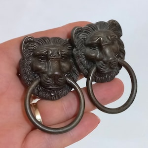 Puede incluir: Dos pomos de puerta antiguos con cabeza de león y tiradores de anillo. Las cabezas de león son de metal y tienen un acabado marrón oscuro.