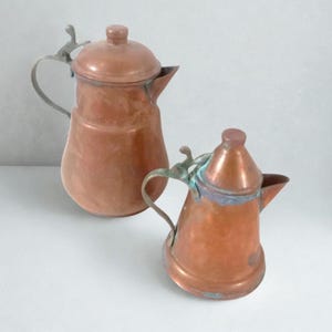 May include: Two copper pitchers with lids and curved metal handles. The larger pitcher has a rounded body and a wide spout, while the smaller pitcher features a tapered top and a spout. Both items show signs of oxidation and patina.
