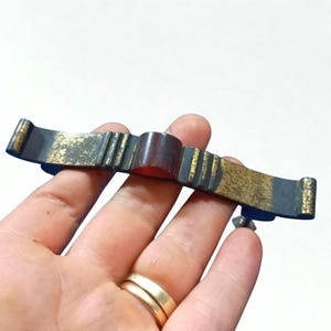 Pode incluir: Um puxador de gaveta vintage em tom bronze com um sotaque central castanho-avermelhado. O puxador retangular apresenta um acabamento desgastado com detalhes dourados e um pequeno parafuso para montagem. O design inclui ranhuras verticais e extremidades curvas.
