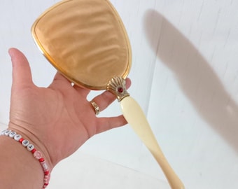 May include: A vintage hand mirror with a gold-toned frame and a cream-colored handle. The mirror has a decorative shell-shaped accent with a red jewel. The mirror's surface has a light, swirling pattern.