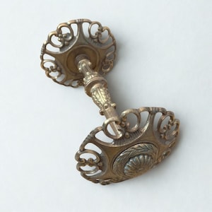 Pode incluir: Uma maçaneta de porta antiga em latão com placas circulares vazadas e ornamentadas. A maçaneta apresenta uma haste central com detalhes decorativos e um motivo floral. O latão tem uma pátina quente e envelhecida, sugerindo charme e uso vintage.