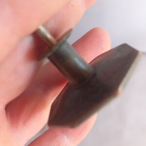 May include: A dark brown, hexagonal-shaped cabinet knob with a smooth, matte finish. The knob features a central cylindrical post that connects to a threaded metal screw extending from the back, designed for attachment to furniture or cabinetry.