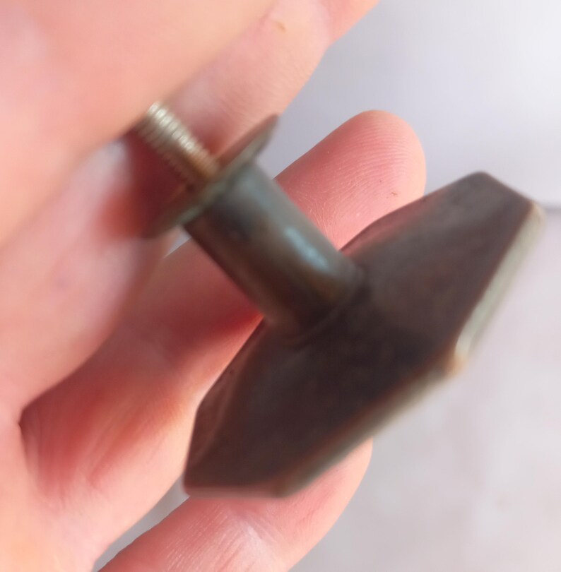 May include: A dark brown, hexagonal-shaped cabinet knob with a smooth, matte finish. The knob features a central cylindrical post that connects to a threaded metal screw extending from the back, designed for attachment to furniture or cabinetry.