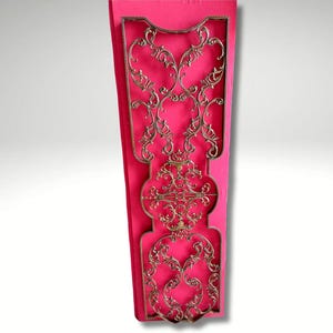 Puede incluir: Un diseño decorativo de filigrana metálica dorada sobre un fondo rosa brillante. La filigrana presenta un patrón ornamental y simétrico con líneas onduladas y elementos florales. El diseño está enmarcado.