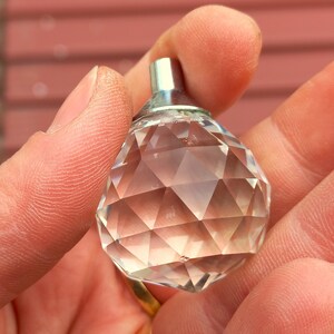 Puede incluir: Un pomo de cristal transparente y facetado con una base de metal plateado. El cristal está tallado con numerosas formas geométricas, que reflejan la luz. El pomo se sostiene en una mano, con un fondo rojo borroso.