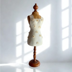 May include: A sleeveless off-white garment featuring a scalloped neckline and scattered small fabric flower appliqués, displayed on a wooden mannequin torso with a matching wooden stand and a rounded base, showcasing a textured surface finish.