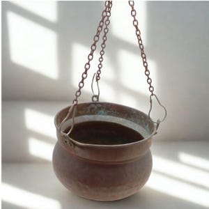 Può includere: Un vaso sospeso color rame con un corpo arrotondato e un bordo largo. Il vaso è sospeso da una catena. Il vaso ha un aspetto vintage e viene probabilmente utilizzato per scopi decorativi.