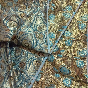 Puede incluir: Primer plano de una tela ornamentada con un patrón floral dorado, turquesa y marrón. La tela tiene un brillo metálico y un diseño texturizado detallado. El patrón incluye flores y hojas estilizadas.