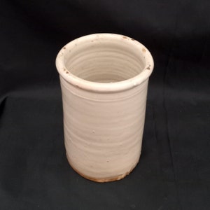 Op de afbeelding: Een cilindrische, crèmekleurige keramische keukengerei houder. De houder heeft een licht getextureerd oppervlak met horizontale ribbels en een afgeronde rand. De basis heeft een lichtbruine tint. De binnenkant is hol en de opening heeft een diameter van ongeveer 10 cm.
