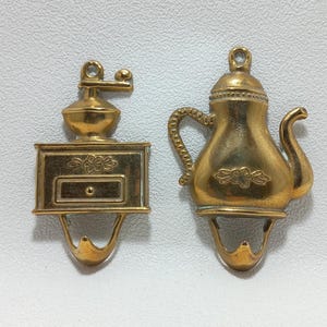 以下が含まれることがあります： 2つのヴィンテージスタイルの真鍮製フック。1つはコーヒーグラインダー、もう1つはティーポットの形をしています。どちらも吊り下げ用のループと湾曲したフックが付いています。コーヒーグラインダーのフックには引き出しと花のディテールがあります。