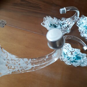 K&ouml;nnte beinhalten: Ein Servierst&uuml;ck aus klarem Glas mit drei gebogenen Armen, die jeweils in einer silbernen Kappe enden. Die Arme haben ein dekoratives, strukturiertes Design mit blauen und wei&szlig;en Akzenten. Auf einer Holzoberfl&auml;che.