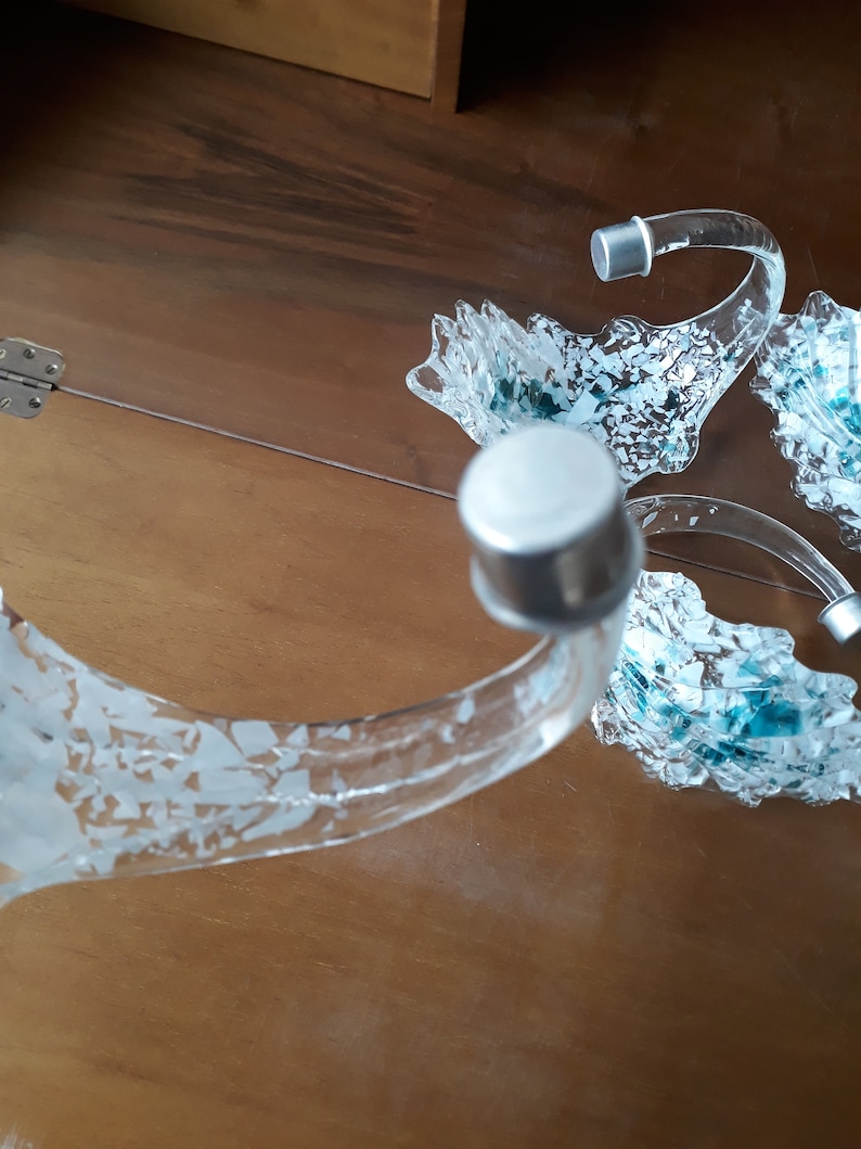 K&ouml;nnte beinhalten: Ein Servierst&uuml;ck aus klarem Glas mit drei gebogenen Armen, die jeweils in einer silbernen Kappe enden. Die Arme haben ein dekoratives, strukturiertes Design mit blauen und wei&szlig;en Akzenten. Auf einer Holzoberfl&auml;che.