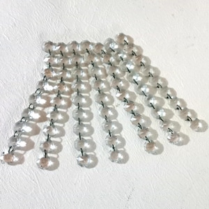 May include: Clear crystal chandelier prisms arranged in a fan shape. Each prism is round and faceted, strung together with thin, dark-colored wire. The prisms catch the light, creating a sparkling effect. Ideal for home decor and lighting projects.