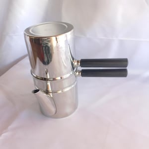 May include: A polished stainless steel stovetop espresso maker featuring a cylindrical body, a small spout, and two horizontal black handles stacked vertically on the side. The metallic surface is reflective and smooth with a clean, simple design.